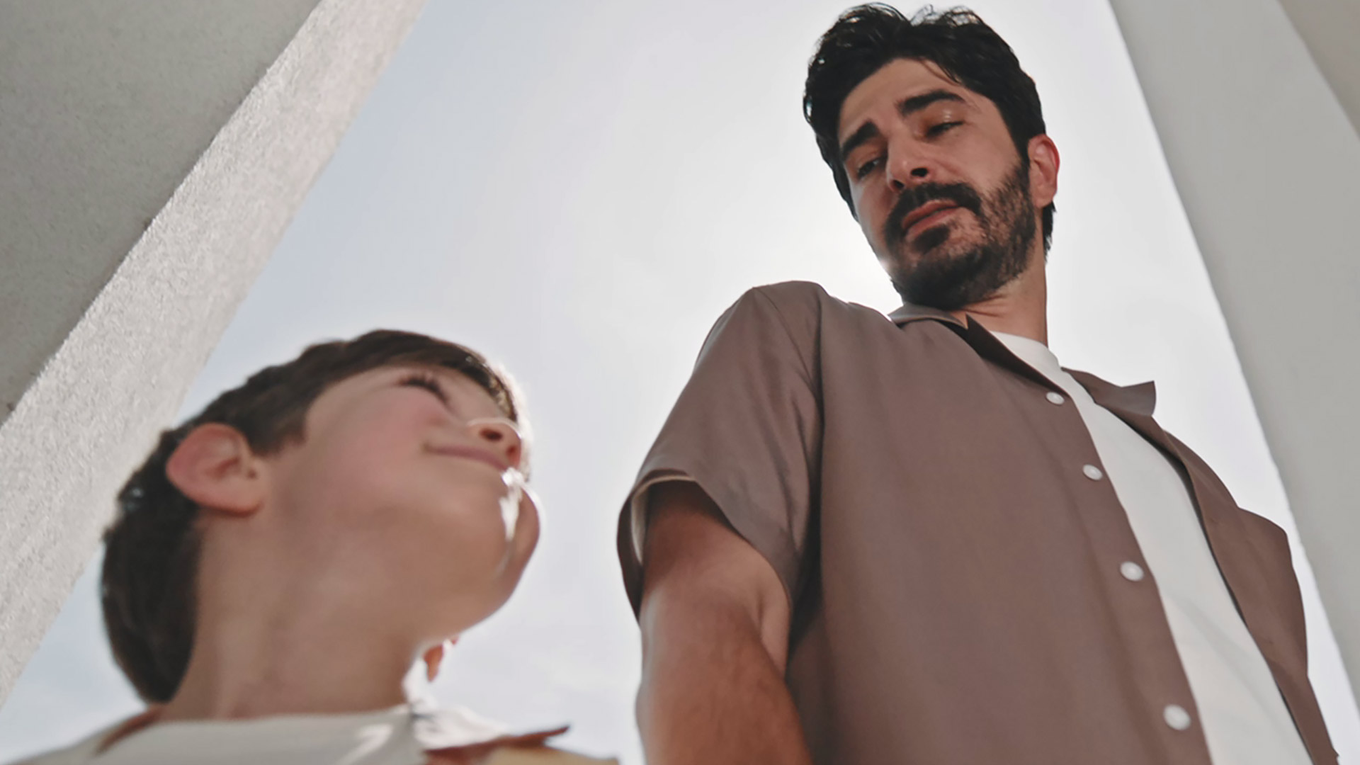 Imagine surprinde un copil și un adult priviți de jos în sus, aflându-se în exterior, cu cerul luminos în fundal. Copilul zâmbește și poartă o cămașă albă, iar adultul este îmbrăcat într-o cămașă maro deschis peste un tricou alb. Coloanele albe ale unei clădiri sunt vizibile pe părțile laterale ale imaginii.
