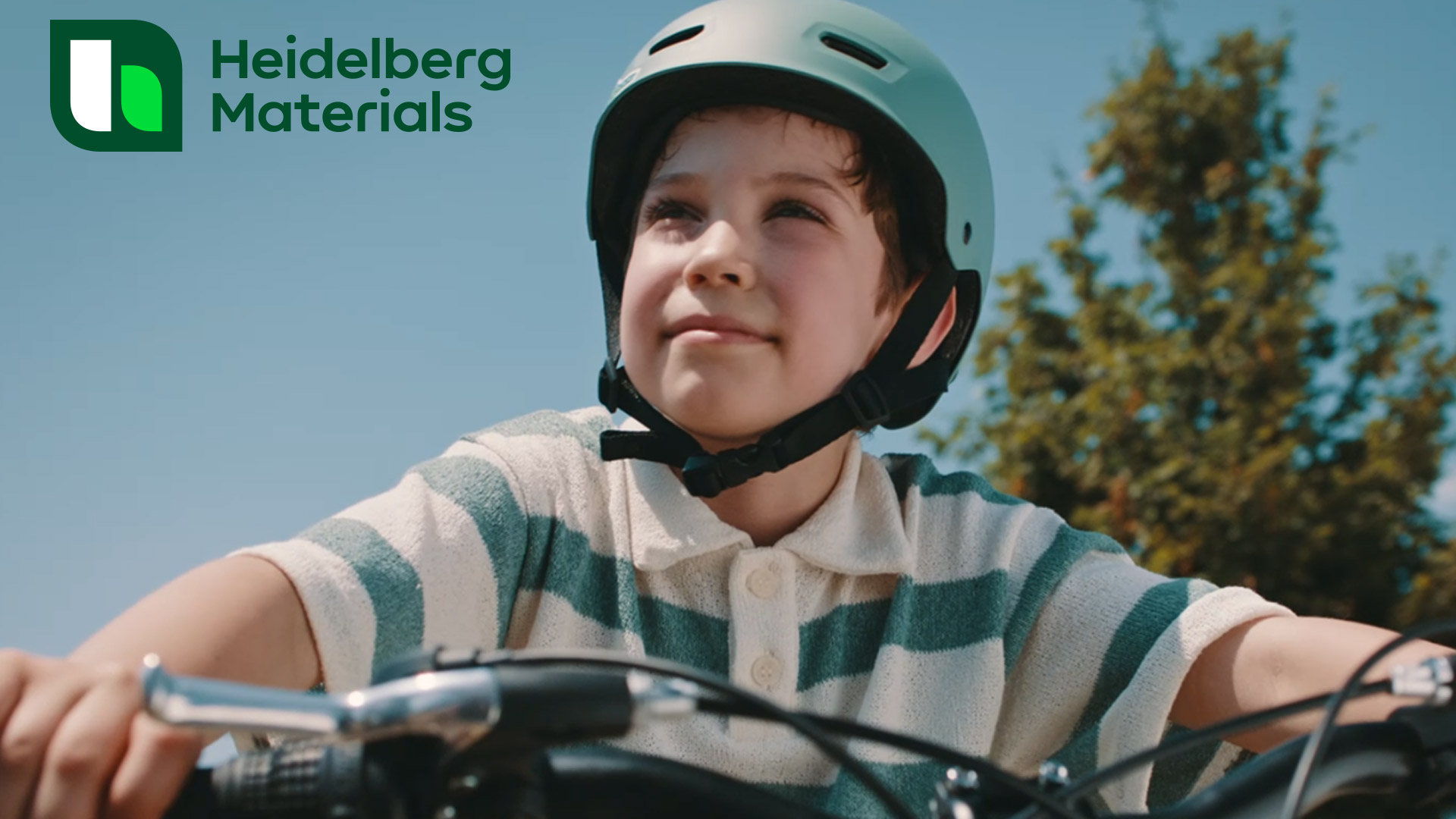 copil pe bicicletă, zâmbind, având pe cap cască de protecție. În colțul din stânga sus este sigla Heidelberg Materials.